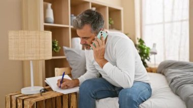 Yakışıklı, beyaz saçlı, ciddi bir telefon konuşmasına dalmış, yatakta oturmuş, notlar alıyor, ders çalışıyor, yatak odasının sıcaklığıyla çevrili.