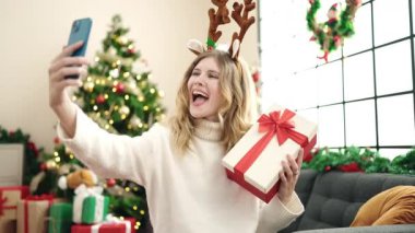 Genç sarışın kadın evde elinde Noel hediyesiyle akıllı telefondan konuşuyor.