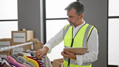 Ciddi, çekici İspanyol genç adam, gri saçlı gönüllü giysileri kontrol ediyor, yardım merkezinin deposundaki pano kontrol listesine not alıyor.