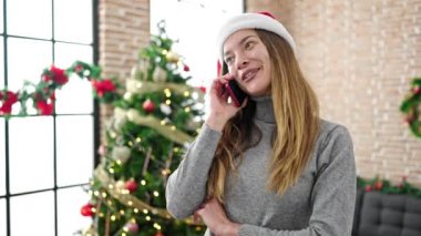 Noel Baba şapkalı mutlu genç kız Noel için dekore edilmiş oturma odasında telefonla konuşuyor.