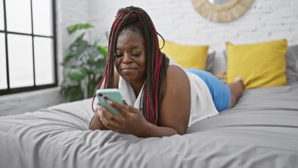 パジャマの美しいアフリカ系アメリカ人女性は 朝のテキストにスマートフォンを使用してベッドでリラックスします 居心地の良い家の雰囲気は彼女の寝室でこの肖像画を組み立てます — ストック動画