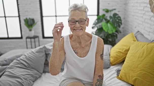 ベッドルームのベッドで快適に座っている間 リラックスした朝のメガネを楽しみながら 灰色の髪の笑顔の自信を持ったシニア女性 — ストック動画