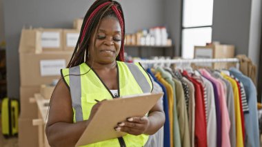Afro-Amerikan kadın, neşeli bir şekilde gülümsüyor, bir hayır kurumunda gönüllü olarak çalışıyor, yansıtıcı yeleğiyle duruyor, bağış kutularıyla çevrili bir pano kontrol listesine yazılmış.