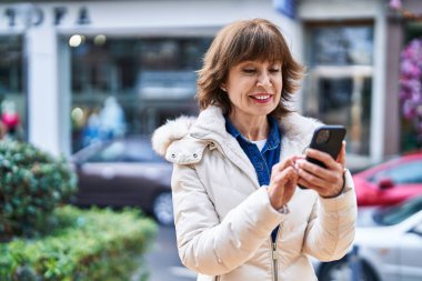 Sokakta akıllı telefon kullanan orta yaşlı bir kadın gülümsüyor.