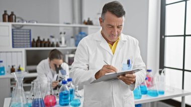 Erkek ve kadın bilim adamları laboratuvarda birlikte çalışıyorlar. Araştırmalarına ciddi şekilde yoğunlaşıyorlar. Mikroskop kullanarak panoya yazı yazıyorlar.