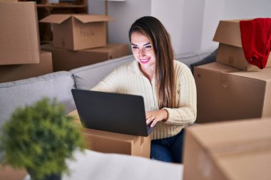 Young beautiful hispanic woman using laptop sitting on sofa at new home