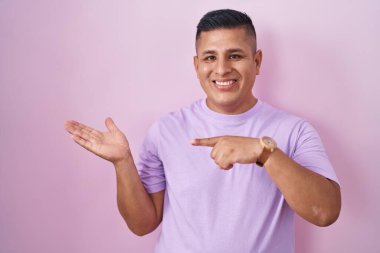 Young hispanic man standing over pink background amazed and smiling to the camera while presenting with hand and pointing with finger. 