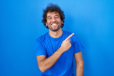 İspanyol asıllı genç adam mavi arka planda neşeli, yüzünde bir gülümseme, mutlu ve doğal bir ifadeyle elini ve parmağını yana kaldırıyor. 