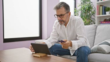 Evdeki kanepede oturan, dokunmatik ped ve kredi kartı kullanan, Latin kökenli, kır saçlı genç bir adam.