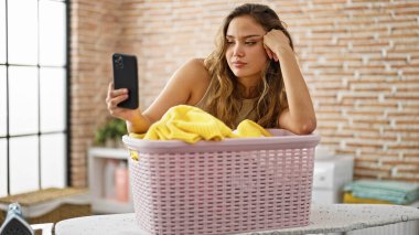 Genç ve güzel İspanyol kadın çamaşır odasında çamaşır sepetine yaslanmış akıllı telefonuyla selfie çekiyor.