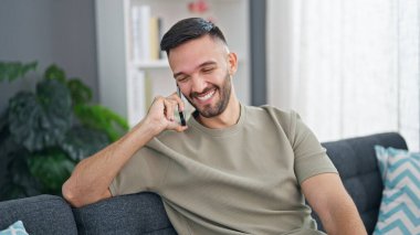 Genç İspanyol adam telefonda konuşuyor. Evdeki kanepede oturuyor.