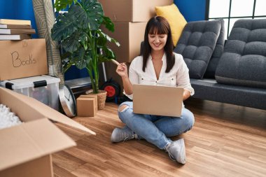 Genç esmer kadın yeni evinde dizüstü bilgisayarla yerde oturuyor gülümsüyor mutlu bir şekilde elini ve parmağını yana doğru gösteriyor. 