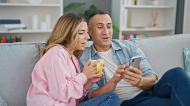 Kadın ve erkek, evdeki kanepede oturmuş akıllı telefon kullanarak kahve içiyorlar.