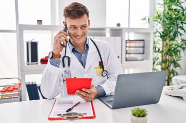 Young man doctor talking on smartphone using touchpad at clinic