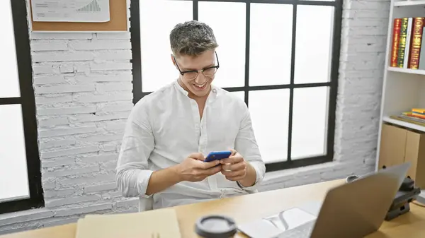 Hardworking young caucasian man in business, beaming while typing a message via smartphone at the office desk.