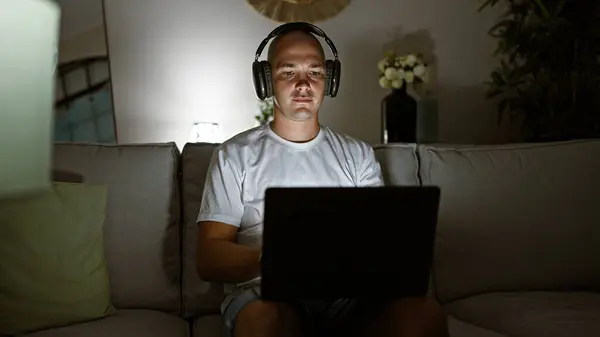 年轻的他的惊慌失措的男人坐在家里沙发上 用笔记本电脑和耳机 — 图库照片