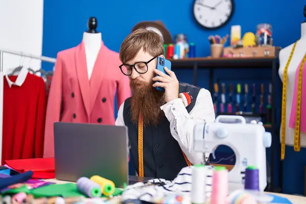 Young redhead man tailor talking on smartphone using laptop at clothing factory