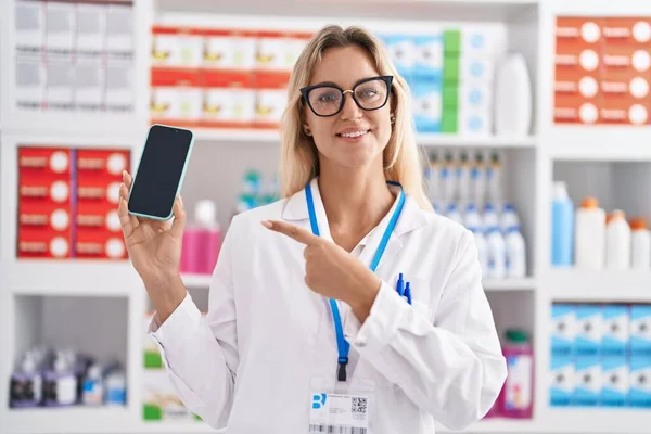 Young Blonde Woman Working Pharmacy Drugstore Showing Smartphone Screen Smiling — Stockfoto