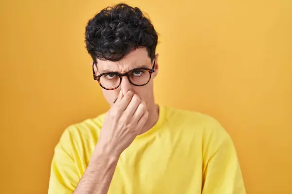 黄色い背景の上に立って眼鏡をかけたヒスパニック男性は 臭いと嫌な臭い 耐え難い匂いを嗅ぎ 鼻に指で息を止めています 悪い匂いがする — ストック写真