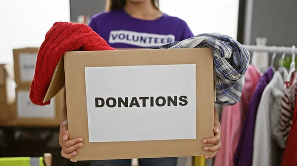Altruistic hands of a community-service driven hispanic woman volunteer, passionately working at the charity center, packing clothes donations into a cardboard box indoors