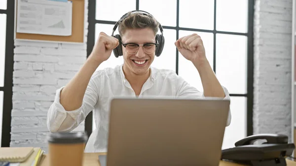 Victorious young caucasian man celebrating winning business success at office, exuding confidence with laptop and headphones amidst festivity