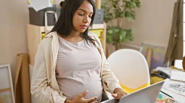 热切的年轻孕妇坐在她的艺术工作室桌旁 精通她的手艺 在画布上画画 用笔记本电脑作为向导 — 图库照片