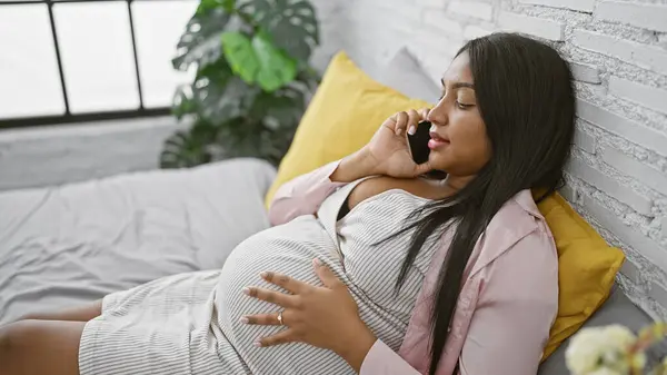 Expecting latin mom relaxed at home, sitting comfortably on bed during morning chatting, touching baby bump in serious pregnancy talk via smartphone, private space in her bedroom interior.