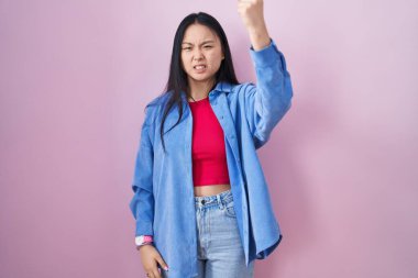 Young asian woman standing over pink background angry and mad raising fist frustrated and furious while shouting with anger. rage and aggressive concept. 