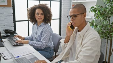 İşteki güçlü çift, iki odaklanmış ofis çalışanı, erkek ve kadın, bilgisayar ve akıllı telefondan ustalıkla çok sayıda iş yapıyorlar.