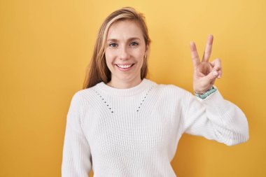 Sarı arka plan üzerine beyaz kazak giyen genç beyaz kadın iki numaralı parmaklarını gösterip gülümserken kendinden emin ve mutlu.. 