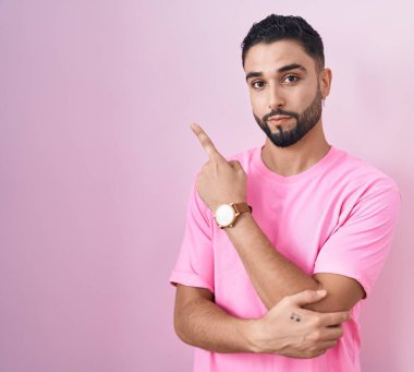 Pembe arka planda duran İspanyol genç adam, elini kenarı işaret ediyor, ciddi ve sakin bir yüz ifadesi sergiliyor. 