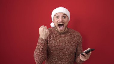 Kafkasyalı genç adam kırmızı arka planda noel şapkası takarak akıllı telefon kullanıyor.