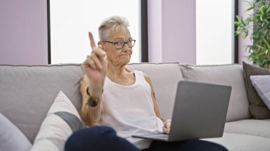 Ciddi, kır saçlı, son sınıf öğrencisi, sıkı bir parmak hareketiyle hayır diyen ve rahat bir ev kanepesinde dizüstü bilgisayar kullanan bir kadın.