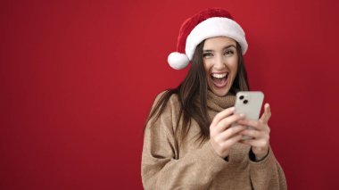 Güzel İspanyol kadın, yılbaşı şapkası takıyor ve izole edilmiş kırmızı arka plan üzerinde akıllı telefon kullanıyor.