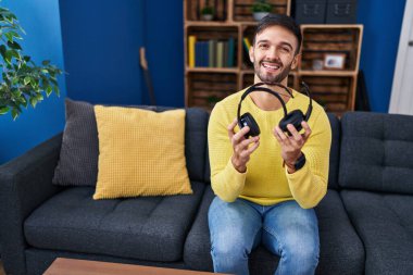 Yüzünde mutlu ve havalı bir gülümsemeyle gülümseyen kırık kulaklık tutan İspanyol bir adam. diş gösterme. 