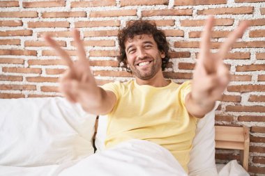 Young hispanic man doing victory gesture sitting on bed at bedroom