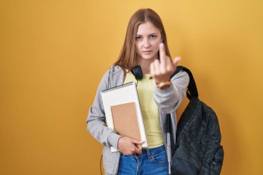 Young caucasian woman wearing student backpack and holding books showing middle finger, impolite and rude fuck off expression 