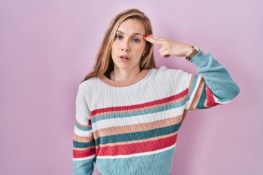 Young blonde woman standing over pink background shooting and killing oneself pointing hand and fingers to head like gun, suicide gesture. 