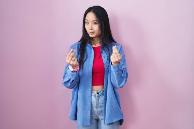 Young asian woman standing over pink background doing money gesture with hands, asking for salary payment, millionaire business 