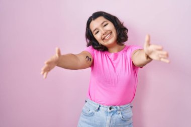 Pembe arka planda duran genç İspanyol kadın kameraya sarılmak için kollarını açıp gülümseyerek bakıyor. Mutluluğu kucaklayan neşeli bir ifade.. 