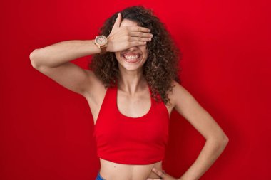 Kıvırcık saçlı İspanyol kadın kırmızı arka planda durup gülümsüyor ve elleri yüzünde şaşkınlıktan gözleri kapayarak gülüyor. kör kavram. 