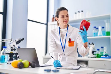 Young beautiful hispanic woman scientist holding pepper writing on document at laboratory