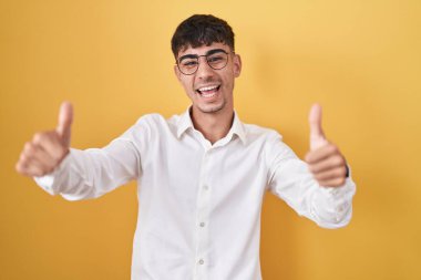 Young hispanic man standing over yellow background approving doing positive gesture with hand, thumbs up smiling and happy for success. winner gesture. 
