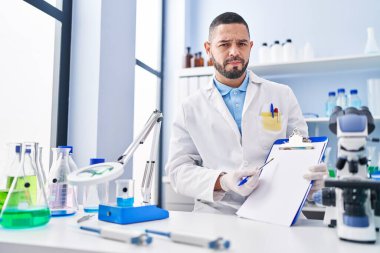 Hispanic man working at scientist laboratory holding blank clipboard skeptic and nervous, frowning upset because of problem. negative person. 