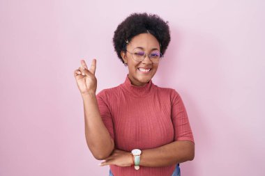 Pembe arka planda duran kıvırcık saçlı güzel Afrikalı kadın kameraya gülümseyen yüzüyle parmaklarıyla zafer işareti yapıyor. İki numara.. 