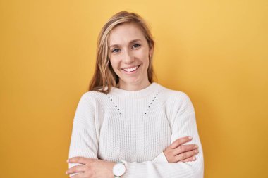 Sarı arka planda beyaz kazak giyen, kollarını kavuşturup kameraya gülümseyen beyaz bir kadın. pozitif kişi. 