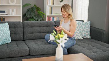 Evdeki kanepede oturan akıllı telefon kullanan genç sarışın kadın.