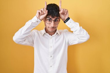 Young hispanic man standing over yellow background doing funny gesture with finger over head as bull horns 