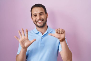 İspanyol adam pembe arka planda duruyor. Altıncı parmaklarını gösterip işaret ediyor. Kendinden emin ve mutlu bir şekilde gülümsüyor.. 