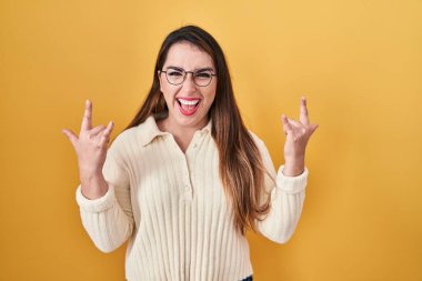 Young hispanic woman standing over yellow background shouting with crazy expression doing rock symbol with hands up. music star. heavy concept. 